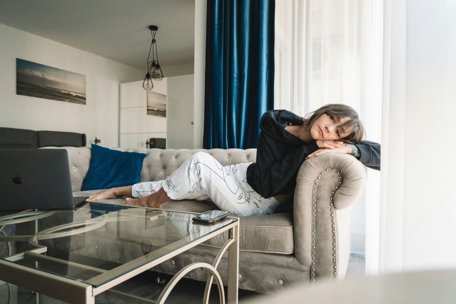 Femme assise en mode détente sur un canapé, vêtue simplement, regard apaisé tourné vers la lumière naturelle.